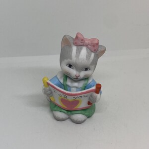 Peut inclure: Figurine en céramique d'un chaton gris et blanc portant un nœud rose, une chemise bleue et une jupe verte. Le chaton tient une bannière avec l'inscription "FOR YOU" et un cœur. La figurine a une expression douce et innocente.