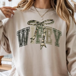 May include: Beige sweatshirt with the word "Wifey" in a distressed, collegiate-style font. The "Wifey" text has a camouflage bow design. The sweatshirt is a neutral color, suitable for casual wear.