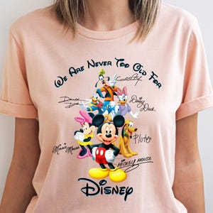 May include: A light pink t-shirt featuring a graphic of Disney characters including Mickey Mouse, Minnie Mouse, Donald Duck, and Goofy. The text "We Are Never Too Old For" and "Disney" are also displayed.