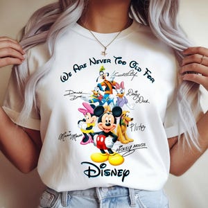 May include: White t-shirt featuring a colorful graphic of Disney characters including Mickey Mouse, Minnie Mouse, Donald Duck, and Goofy. The text "We Are Never Too Old For Disney" is printed above the characters.