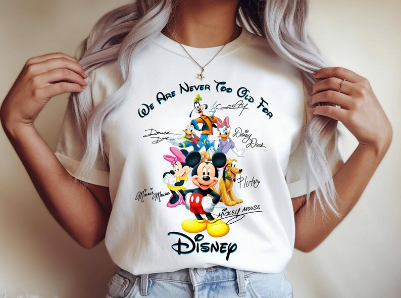 May include: White t-shirt featuring a colorful graphic of Disney characters including Mickey Mouse, Minnie Mouse, Donald Duck, and Goofy. The text "We Are Never Too Old For Disney" is printed above the characters.