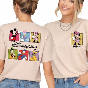 May include: Beige t-shirts featuring Disney characters. One shirt has a Disneyland graphic with Mickey Mouse, Donald Duck, and Goofy. The other shirt has a Minnie Mouse graphic with the word "MOM".