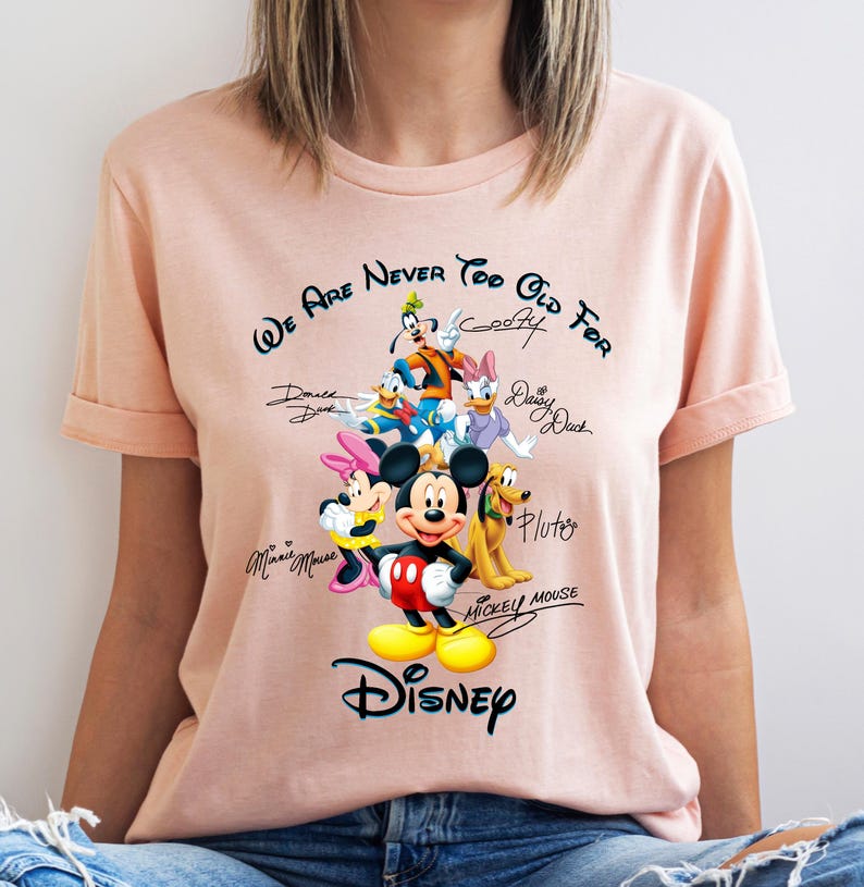 May include: A light pink t-shirt featuring a graphic of Disney characters including Mickey Mouse, Minnie Mouse, Donald Duck, and Goofy. The text "We Are Never Too Old For" and "Disney" are also displayed.