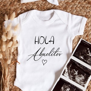 May include: White onesie with the text "Hola Abuelitos" in black lettering with a small heart. Three black and white ultrasound images are on a woven surface.