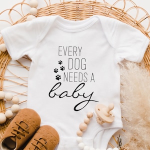 May include: White baby onesie with black text that reads "Every dog needs a baby". The onesie is lying on a woven wicker surface with a wooden bead necklace and a pair of brown baby shoes.