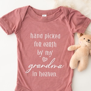 May include: A dusty rose baby onesie with the text "hand picked for earth by my grandma in heaven."  A small teddy bear is in the background.