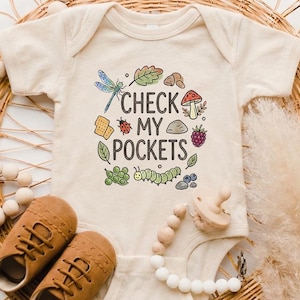 May include: A cream-colored baby onesie with the text "CHECK MY POCKETS" and colorful illustrations of a dragonfly, leaves, a ladybug, and other nature elements. Brown baby shoes and a pacifier are also visible.
