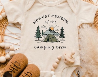 May include: A cream-colored short-sleeve baby bodysuit featuring a graphic of a mountain, a tent, and pine trees with the text "NEWEST MEMBER of the camping crew" printed in black. The design includes a small orange sun above the mountain.