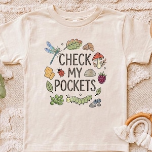 May include: Cream-colored t-shirt with a colorful graphic design. The design includes a dragonfly, leaves, a mushroom, a ladybug, a caterpillar, and the text "CHECK MY POCKETS".