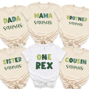 May include: A collection of beige and white t-shirts with dinosaur-themed text. The shirts say "DADA saurus", "MAMA saurus", "BROTHER saurus", "SISTER saurus", "ONE REX", and "COUSIN saurus" in green and black lettering.