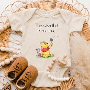 A baby onesie with a Winnie the Pooh character, brown shoes, and a beaded necklace are arranged on a woven basket.