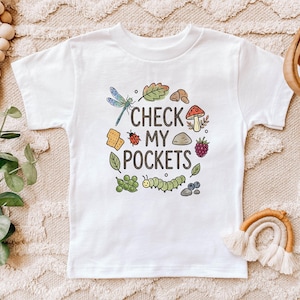 May include: White t-shirt with the text "CHECK MY POCKETS" surrounded by colorful illustrations of a dragonfly, mushroom, caterpillar, and other nature-themed elements. The shirt is a children's size.