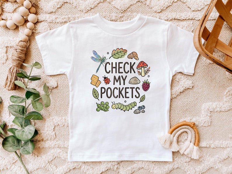 May include: White t-shirt with the text "CHECK MY POCKETS" surrounded by colorful illustrations of a dragonfly, mushroom, caterpillar, and other nature-themed elements. The shirt is a children's size.