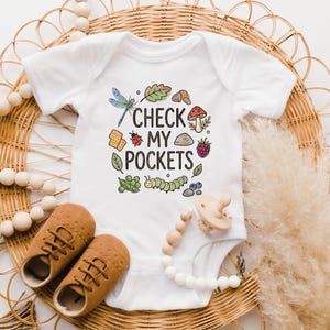 May include: White baby onesie with the text "CHECK MY POCKETS" and colorful illustrations of a dragonfly, leaf, mushroom, ladybug, and other nature elements. Brown baby shoes and a pacifier are also in the image.