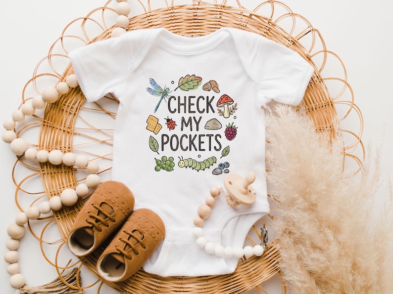 May include: White baby onesie with the text "CHECK MY POCKETS" and colorful illustrations of a dragonfly, leaf, mushroom, ladybug, and other nature elements. Brown baby shoes and a pacifier are also in the image.