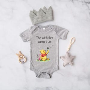 A gray onesie with a Winnie the Pooh character, a gray crown, a wooden bead rattle, and a gray star are arranged on a marble surface.