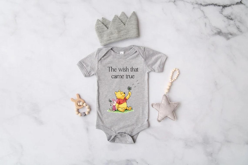 A gray onesie with a Winnie the Pooh character, a gray crown, a wooden bead rattle, and a gray star are arranged on a marble surface.