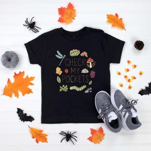 May include: Black t-shirt with the text "CHECK MY POCKETS" surrounded by a colorful illustration of a dragonfly, leaves, and other nature elements. Gray sneakers and fall-themed decorations surround the shirt.