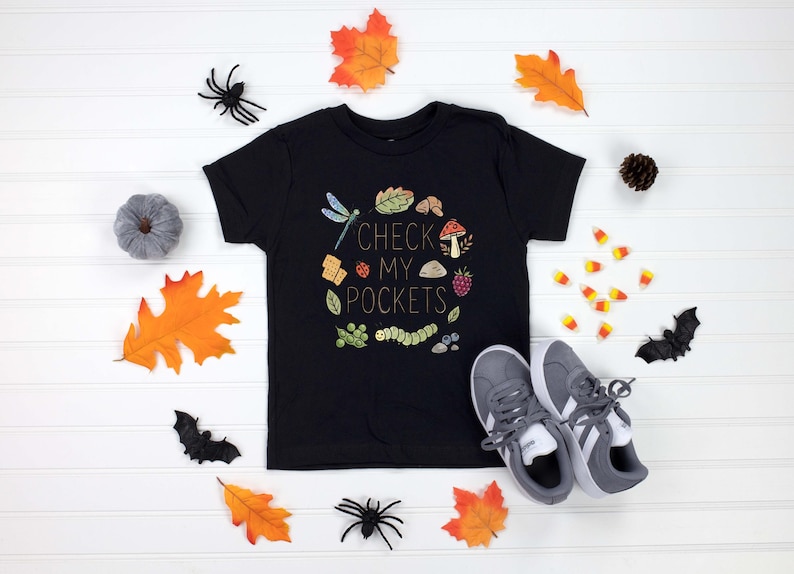 May include: Black t-shirt with the text "CHECK MY POCKETS" surrounded by a colorful illustration of a dragonfly, leaves, and other nature elements. Gray sneakers and fall-themed decorations surround the shirt.