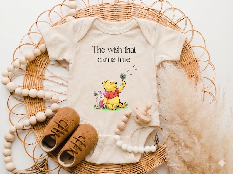 A baby onesie with a Winnie the Pooh character, brown shoes, and a beaded necklace are arranged on a woven basket.