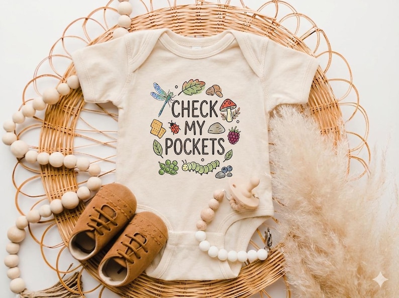 May include: A cream-colored baby onesie with the text "CHECK MY POCKETS" and colorful illustrations of a dragonfly, leaves, a ladybug, and other nature elements. Brown baby shoes and a pacifier are also visible.