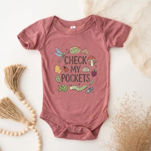 May include: A dusty rose baby bodysuit with a whimsical design. The graphic includes a dragonfly, leaves, a mushroom, and the text "CHECK MY POCKETS". The design is surrounded by other small illustrations. The bodysuit has short sleeves and snap closures.