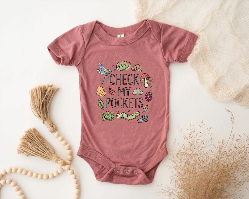 May include: A dusty rose baby bodysuit with a whimsical design. The graphic includes a dragonfly, leaves, a mushroom, and the text "CHECK MY POCKETS". The design is surrounded by other small illustrations. The bodysuit has short sleeves and snap closures.