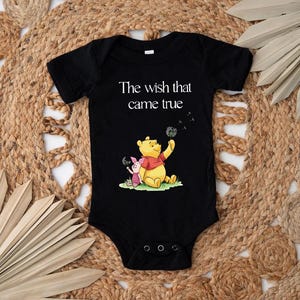 A black baby onesie with a Winnie the Pooh and Piglet graphic, along with the text "The wish that came true" printed on it, is displayed on a woven mat.