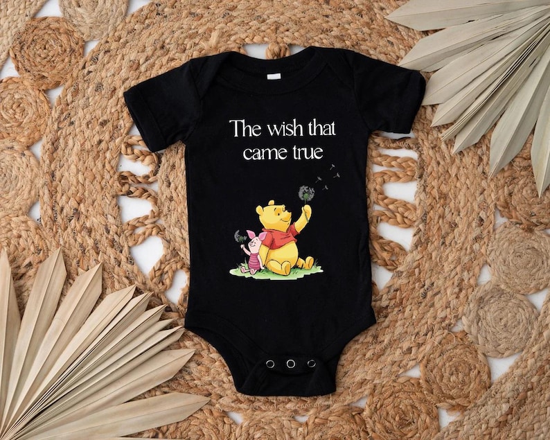 A black baby onesie with a Winnie the Pooh and Piglet graphic, along with the text "The wish that came true" printed on it, is displayed on a woven mat.