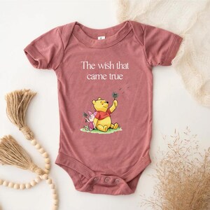 A pink baby onesie with a Winnie the Pooh character printed on it, along with a tassel and some dried plants.