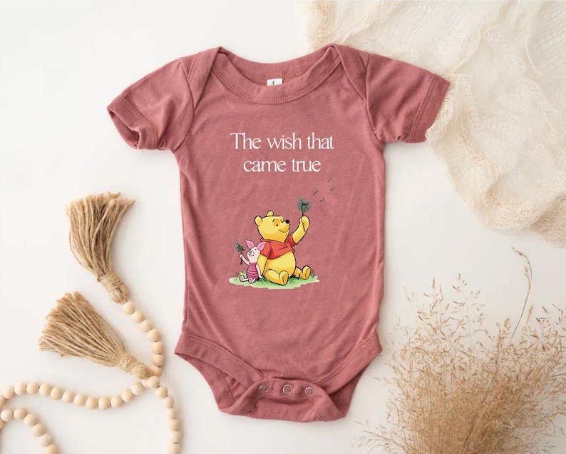 A pink baby onesie with a Winnie the Pooh character printed on it, along with a tassel and some dried plants.