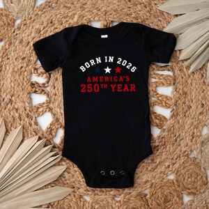 May include: A black short-sleeved baby bodysuit featuring the white text BORN IN 2026, three stars in blue, white, and red, and the red text AMERICA'S 250TH YEAR. The garment has a round neckline and three metal snaps at the bottom crotch area.