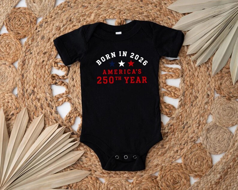 May include: A black short-sleeved baby bodysuit featuring the white text BORN IN 2026, three stars in blue, white, and red, and the red text AMERICA'S 250TH YEAR. The garment has a round neckline and three metal snaps at the bottom crotch area.