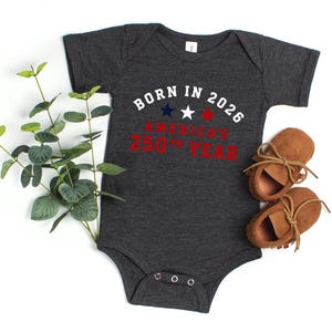 May include: A dark charcoal gray short-sleeve baby bodysuit featuring the white text BORN IN 2026 with three small stars in blue, white, and red, followed by the red text AMERICA'S 250TH YEAR, accompanied by a pair of tan suede baby moccasins.
