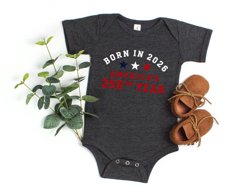May include: A dark charcoal gray short-sleeve baby bodysuit featuring the white text BORN IN 2026 with three small stars in blue, white, and red, followed by the red text AMERICA'S 250TH YEAR, accompanied by a pair of tan suede baby moccasins.