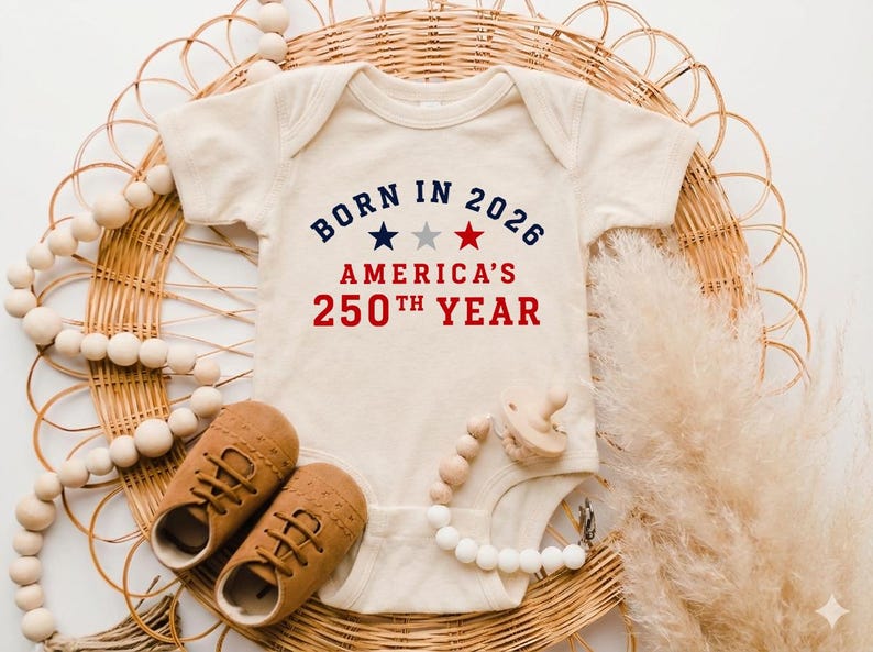 May include: A cream-colored short-sleeve baby bodysuit featuring the text BORN IN 2026 AMERICA'S 250TH YEAR in navy blue and red, with three small stars in navy, gray, and red centered between the text lines. The garment has a round neckline.