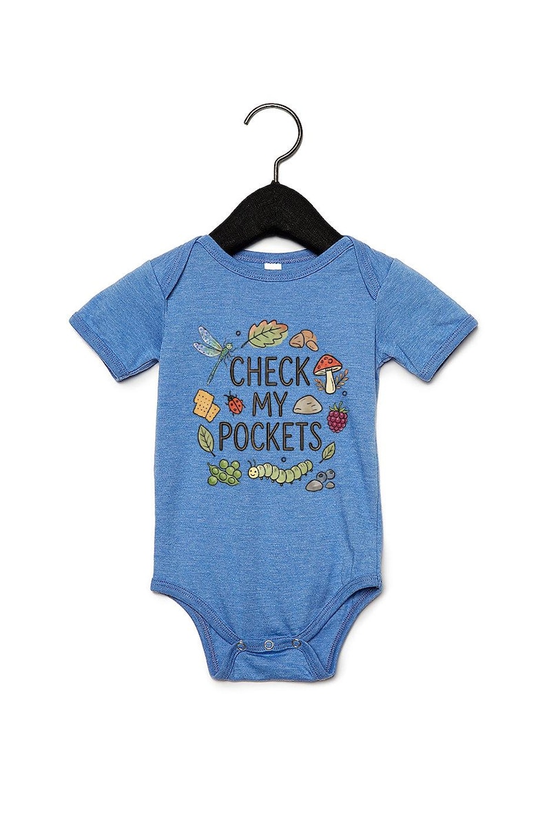 May include: Blue baby bodysuit with the text "CHECK MY POCKETS" and illustrations of a dragonfly, leaves, mushrooms, berries, and a caterpillar. The bodysuit is hanging on a black hanger.