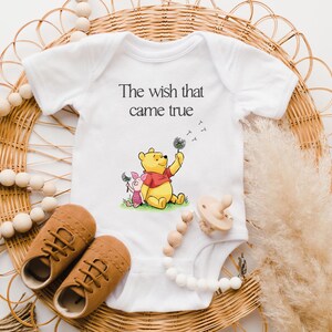 A baby onesie with a Winnie the Pooh character, brown shoes, and a pacifier are arranged on a woven basket, along with some beaded accessories.