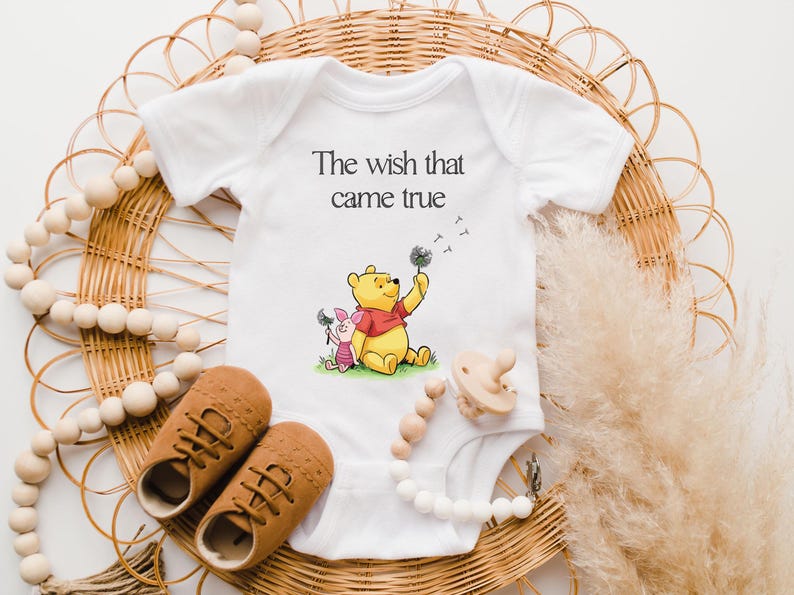 A baby onesie with a Winnie the Pooh character, brown shoes, and a pacifier are arranged on a woven basket, along with some beaded accessories.