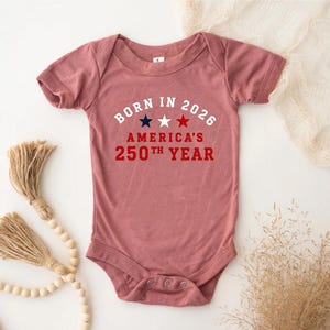 May include: A dusty rose short-sleeve baby bodysuit featuring the white text BORN IN 2026, three stars in navy, white, and red, and the red text AMERICA'S 250TH YEAR. The garment has a crew neckline and a snap closure at the bottom for easy access.