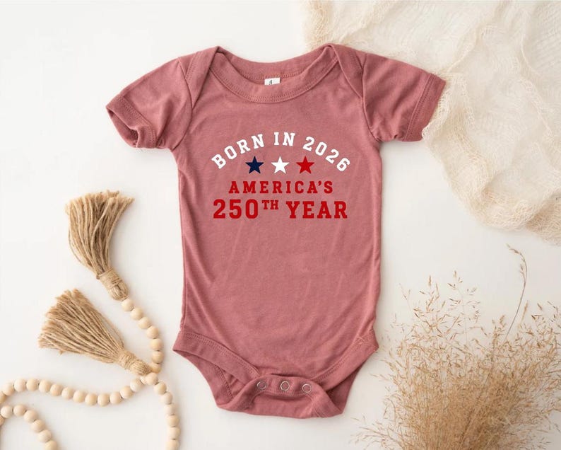 May include: A dusty rose short-sleeve baby bodysuit featuring the white text BORN IN 2026, three stars in navy, white, and red, and the red text AMERICA'S 250TH YEAR. The garment has a crew neckline and a snap closure at the bottom for easy access.