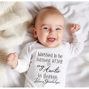 May include: A white baby onesie with black text that reads "blessed to be named after my Auntie in Heaven. Liliana Guadalupe". A small heart symbol is between "my" and "Auntie". A baby is wearing the onesie and smiling.