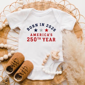 May include: A white short-sleeved baby bodysuit featuring the text BORN IN 2026 AMERICA'S 250TH YEAR in blue and red, with three small stars in blue, grey, and red centered between the lines of text, accompanied by tan shoes and a pacifier.