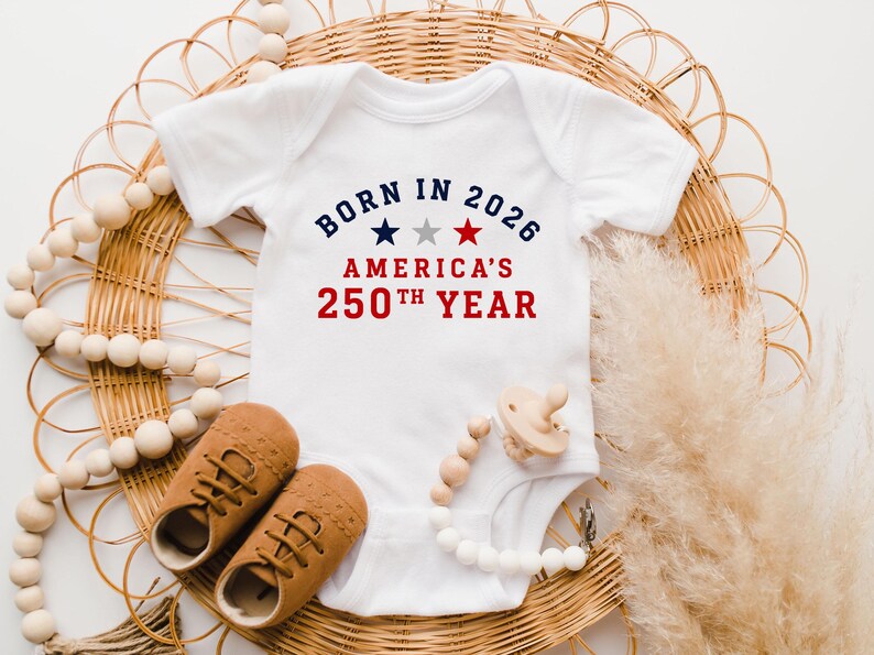 May include: A white short-sleeved baby bodysuit featuring the text BORN IN 2026 AMERICA'S 250TH YEAR in blue and red, with three small stars in blue, grey, and red centered between the lines of text, accompanied by tan shoes and a pacifier.