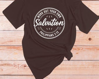 May include: A dark brown t-shirt featuring a white circular design. The design includes the words "Work Out Your Own Salvation" and "Philippians 2:12". The t-shirt is knotted at the hem.