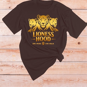 Puede incluir: Una camiseta marrón con un gráfico dorado de tres leonas y las palabras "Lioness Hood" con las frases "One Pride" y "One Roar" debajo. La camiseta está anudada en la parte inferior.
