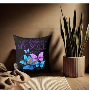 Op de afbeelding: Een zwart kussen met de tekst "He Restores My Soul" in paarse letters en de tekst "Psalm 23:3" in kleinere letters. Het kussen heeft een ontwerp met kleurrijke vlinders.