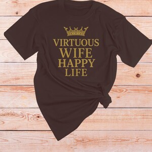 May include: A dark brown t-shirt featuring the gold text "VIRTUOUS WIFE HAPPY LIFE" and a gold crown illustration. The t-shirt is knotted at the bottom.