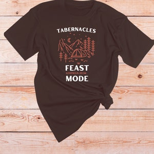 Może przedstawiać: Brązowy T-shirt z graficznym wzorem przedstawiającym scenę górską z namiotem, drzewami i gwiazdami. Na koszulce nadrukowany jest tekst "Tabernacles Feast Leviticus 23:34 Mode".