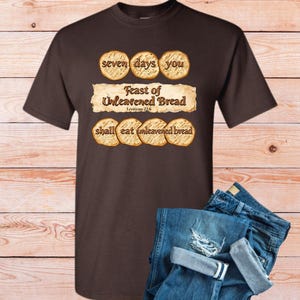 Op de afbeelding: Een bruin t-shirt met de tekst "seven days you Feast of Unleavened Bread shall eat unleavened bread" in een decoratief lettertype. De outfit wordt gecompleteerd door een blauwe jeans met distressed details.